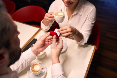 Photo for Cropped photo of guy presenting engagement ring to happy female companion at cafe table - Royalty Free Image