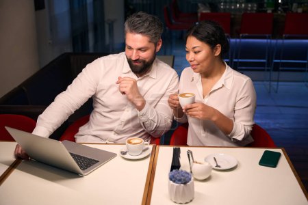 Photo for Smiling young woman and man seated at cafe table looking at laptop screen - Royalty Free Image