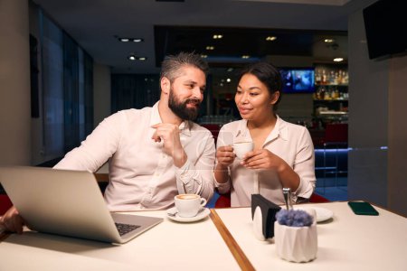 Foto de Hombre alegre sentado en la mesa de la cafetería mostrando algo a su compañera femenina complacida en la pantalla del ordenador portátil - Imagen libre de derechos