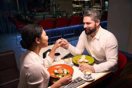 Foto de Sonriente chico complacido y su feliz acompañante femenino tomados de la mano en la mesa del restaurante - Imagen libre de derechos