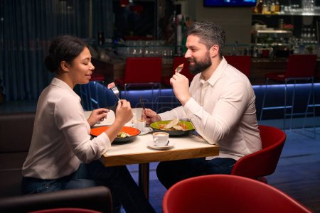 Téléchargez les photos : Agréable jeune femme et son compagnon joyeux manger de la salade à la table du restaurant - en image libre de droit