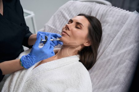 Téléchargez les photos : Femme en manteau médical et gants tenant la seringue et faisant des injections de beauté pour le visage au client - en image libre de droit