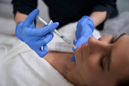 Téléchargez les photos : Femme en manteau médical et gants tenant la seringue et faisant des injections de beauté pour le visage au client - en image libre de droit