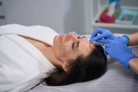 Photo for Young woman lying on couch in the beauty salon, doctor in protective gloves standing near and doing beauty injections - Royalty Free Image