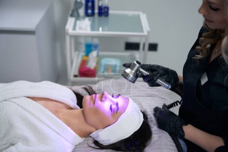 Photo for Cosmetologist in protective gloves sitting near adult woman and doing led therapy procedure in office - Royalty Free Image