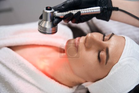 Photo for Cosmetologist in protective gloves sitting near woman and doing led therapy procedure in office - Royalty Free Image