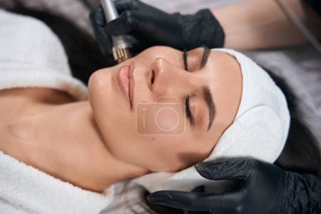 Téléchargez les photos : Cosmetologue dans des gants de protection assis près de dame adulte et faire une procédure d'électroporation pour le visage dans le salon - en image libre de droit