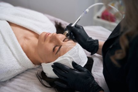 Téléchargez les photos : Cosmetologue dans des gants de protection assis près d'une femme adulte et faisant une procédure d'électroporation pour le visage dans le salon - en image libre de droit