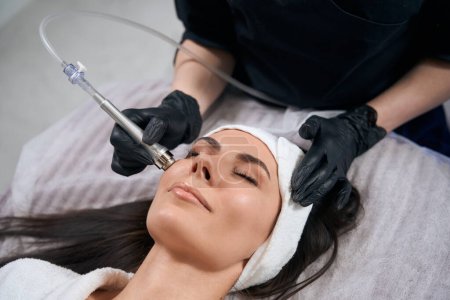 Téléchargez les photos : Cosmetologue dans des gants de protection assis près de belle femme et faire une procédure d'électroporation pour le visage dans le salon - en image libre de droit