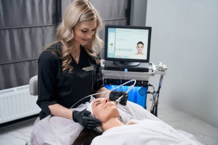 Photo for Cosmetologist in protective gloves sitting near beautiful female and doing electroporation procedure for face in salon - Royalty Free Image