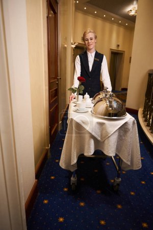 Téléchargez les photos : Jeune serveuse sur un chariot transporte de la nourriture chaude le long du couloir de l'hôtel, une femme en uniforme - en image libre de droit