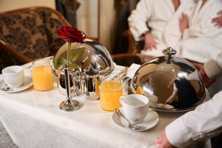 Photo for Hotel waitress serves breakfast in the room for a couple in love, the couple are sitting in bathrobes - Royalty Free Image