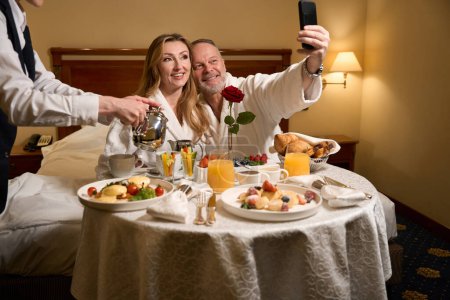 Téléchargez les photos : Couple d'adultes célébrant leur anniversaire de mariage dans un hôtel de luxe, le serveur mis en place un repas romantique pour eux - en image libre de droit