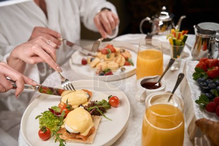 Téléchargez les photos : Homme et femme prenant un petit déjeuner romantique dans une chambre d'hôtel, les conjoints mangent des œufs pochés et des légumes - en image libre de droit