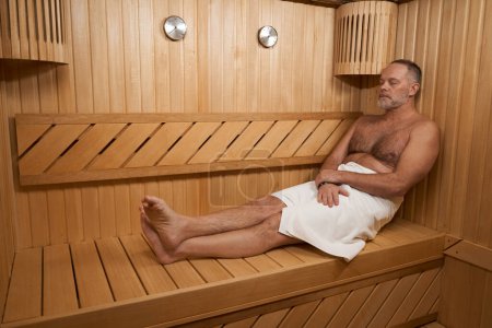 Téléchargez les photos : Homme d'âge moyen avec un torse nu se réchauffe dans un sauna, la chambre est décorée avec du bois - en image libre de droit