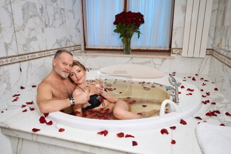Téléchargez les photos : Heureux couple célébrant leur anniversaire de mariage dans un hôtel de luxe, le couple se luxuriera dans un bain aux pétales de rose et champagne - en image libre de droit