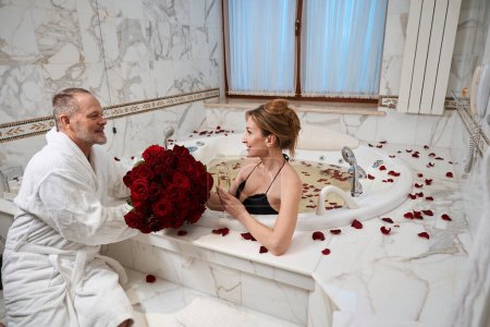 Téléchargez les photos : Homme barbu en peignoir donne bouquet à sa charmante femme, dame éclaboussures dans la salle de bain avec une coupe de champagne - en image libre de droit