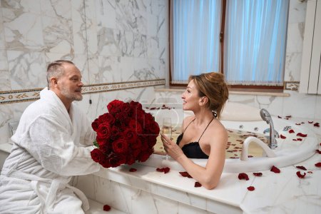 Téléchargez les photos : Homme d'âge moyen donne un bouquet à sa charmante femme, une dame éclabousse dans la salle de bain avec une coupe de champagne - en image libre de droit