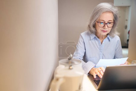 Photo for Lady with glasses is located with a laptop at the kitchen table, she comfortably studies at distance courses - Royalty Free Image