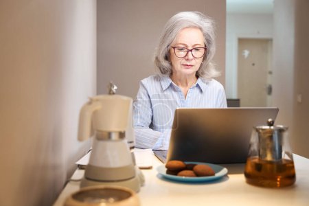 Foto de Hermosa anciana trabaja con un ordenador portátil en la mesa de la cocina, en las galletas de mesa y gadgets de cocina - Imagen libre de derechos