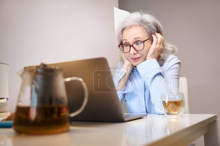 Foto de Jubilado jubiloso se sienta en un ordenador portátil en la mesa de la cocina, en la mesa todo para una fiesta de té - Imagen libre de derechos