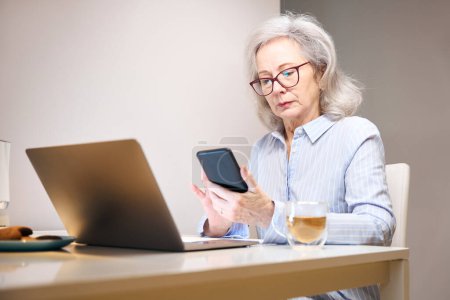 Téléchargez les photos : Dame sérieuse avec des lunettes s'installe avec un ordinateur portable et un téléphone à la table de la cuisine - en image libre de droit