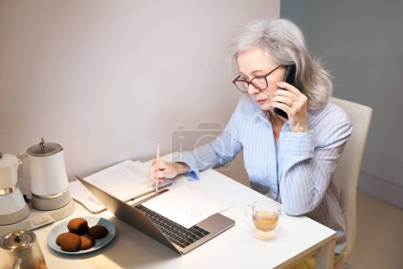 Foto de Serious anciana se sienta en la mesa de la cocina y trabaja con el ordenador portátil y el teléfono, galletas y aparatos de cocina en la mesa - Imagen libre de derechos