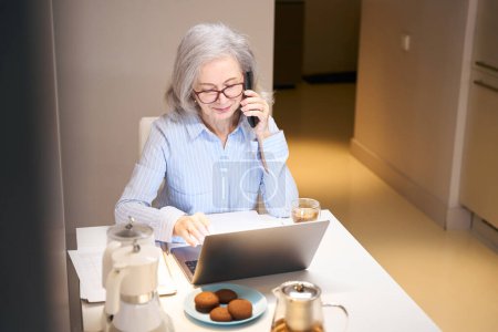 Foto de Sonriendo anciana se comunica tranquilamente por teléfono, ella se instala en la cocina con un ordenador portátil - Imagen libre de derechos