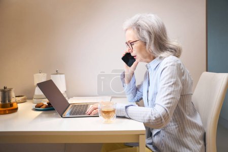 Foto de Señora de edad avanzada moderna trabaja desde casa, se comunica por teléfono, en la mesa de la cocina portátil - Imagen libre de derechos