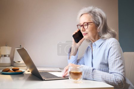 Photo for Active elderly lady works from home, she communicates on the phone, on the kitchen table laptop - Royalty Free Image