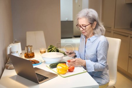 Foto de Mujer de pelo gris en vasos corta verduras para una ensalada saludable, frente a ella en la nota de la mesa de la cocina - Imagen libre de derechos