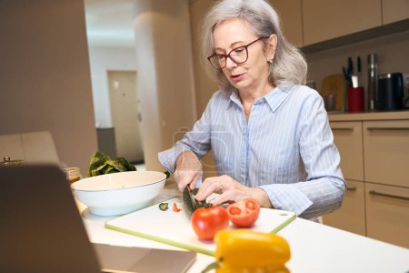 Foto de Pensionista de pelo gris en ropa casual corta verduras para una ensalada saludable, frente a ella en la nota de la mesa de la cocina - Imagen libre de derechos
