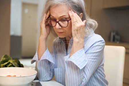Foto de Solitaria anciana se sienta en la mesa de la cocina, sufre de dolor de cabeza - Imagen libre de derechos