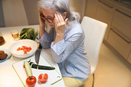 Foto de La anciana se aferra a sus sienes, sufre de dolor de cabeza, se sienta en la mesa de la cocina - Imagen libre de derechos
