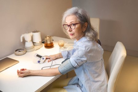 Photo for Pretty elderly lady measures her blood pressure with a tonometer, she is alone sitting at home in the kitchen - Royalty Free Image