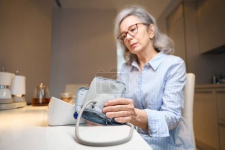 Téléchargez les photos : Femme âgée fatiguée utilise un tonomètre pour surveiller la pression artérielle, elle est assise à la maison dans la cuisine - en image libre de droit