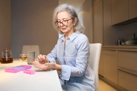 Foto de Mujer mayor se sienta en la cocina y pone pastillas en una caja de pastillas, tiene el pelo gris - Imagen libre de derechos