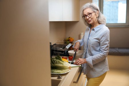 Téléchargez les photos : Agréable femme âgée dans la cuisine à la maison est engagée dans la cuisine, elle coupe des légumes pour une salade - en image libre de droit