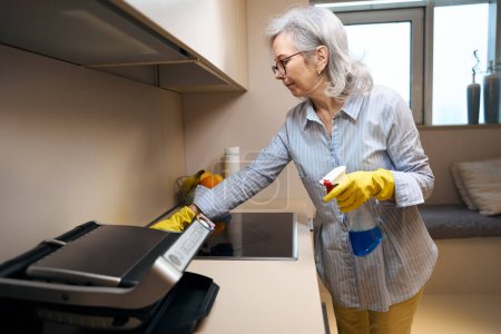 Foto de Mujer de edad limpia lava la cocina con un paño suave, ella utiliza un limpiador especial - Imagen libre de derechos
