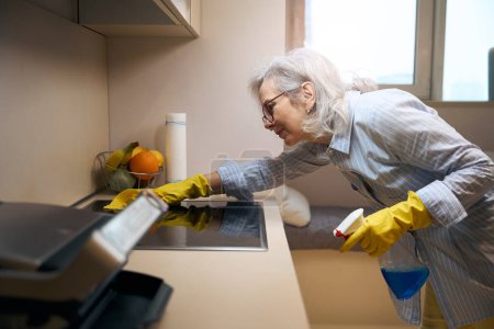 Photo for Neat pensioner washes the kitchen stove with a soft cloth, she uses a special cleaner - Royalty Free Image