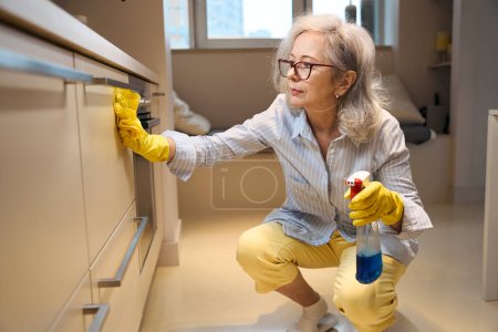 Téléchargez les photos : Femme âgée mince lave les surfaces de cuisine accroupi, elle travaille dans des gants de protection - en image libre de droit