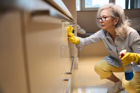 Foto de Linda abuela delgada lava las superficies de la cocina en cuclillas, ella trabaja en guantes de protección - Imagen libre de derechos