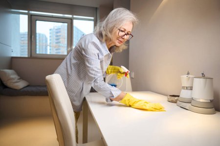 Téléchargez les photos : Mignonne grand-mère en lunettes et gants de protection lave les surfaces de cuisine, elle est en vêtements décontractés - en image libre de droit