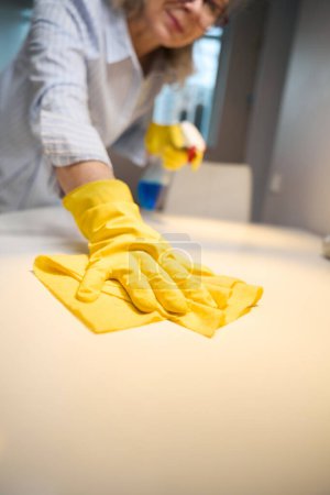 Foto de Mujer mayor lava la mesa de la cocina con un paño suave, ella utiliza un limpiador especial - Imagen libre de derechos