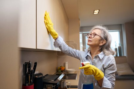 Téléchargez les photos : Grand-mère intelligente dans des lunettes et des gants de protection lave l'armoire de cuisine, elle est en vêtements décontractés - en image libre de droit