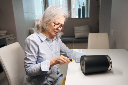 Foto de Mujer avanzada de edad en gafas está reparando un hervidor eléctrico con un destornillador, que se encuentra en la mesa de la cocina - Imagen libre de derechos