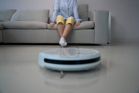 Téléchargez les photos : Femme âgée solitaire assis sur un canapé doux dans le salon, robot aspirateur nettoie le sol dans la chambre - en image libre de droit