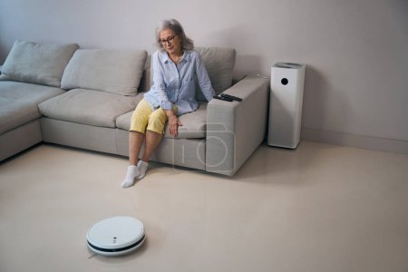Téléchargez les photos : Femme retraitée assise sur le canapé et regarde le travail de l'aspirateur robot, il y a un purificateur d'air à proximité - en image libre de droit