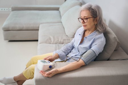Foto de Mujer mayor se sienta en un sofá y mide la presión arterial con un tonómetro, tiene el pelo gris - Imagen libre de derechos