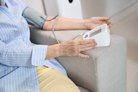 Foto de La mujer se sienta en un sofá y mide la presión arterial con un tonómetro, ella no está bien - Imagen libre de derechos